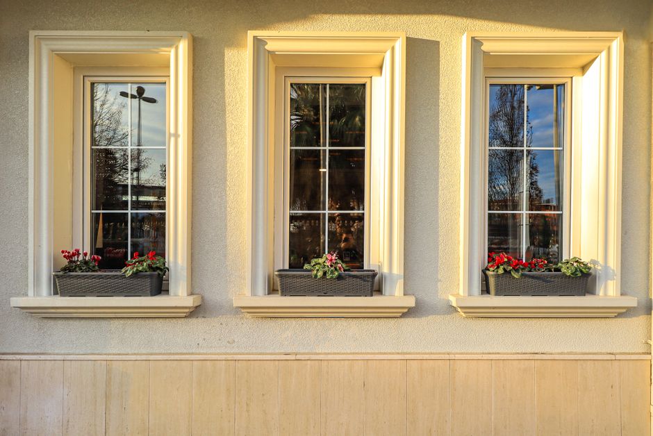 Three tall windows with white trim and window boxes of red flowers on a pale stucco wall in low sunlight