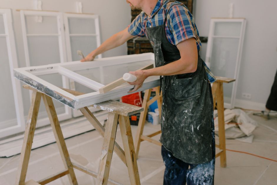 Worker in a paint-spattered apron sanding a white window sash laid across two sawhorses