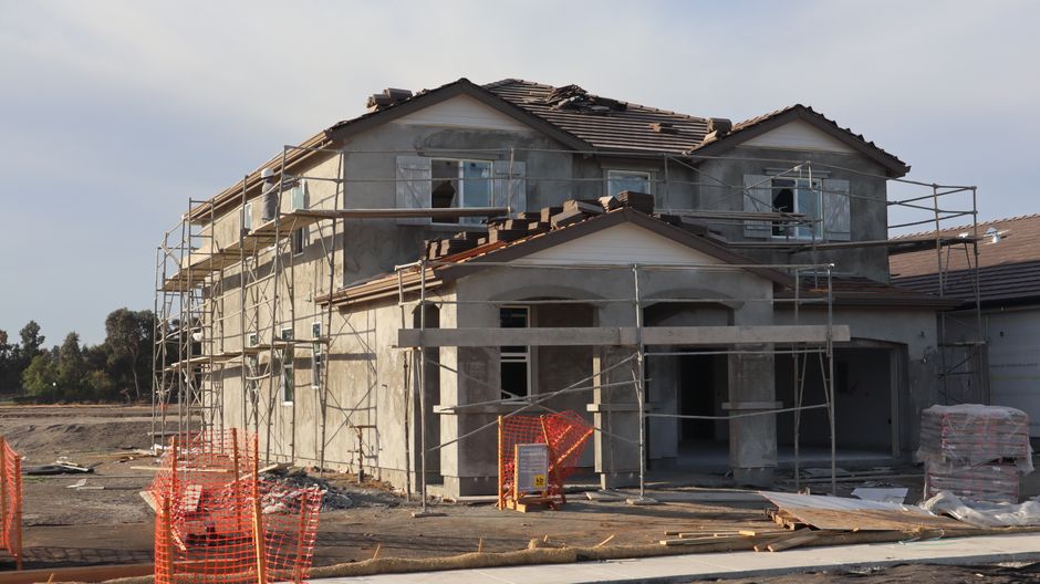 Two-story stucco house under construction with scaffolding, stacked roof tiles and orange safety fencing in front