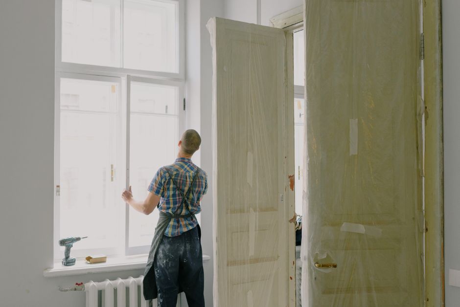 Installer in an apron standing at a tall white window in a room with plastic-wrapped doors and a drill on the sill