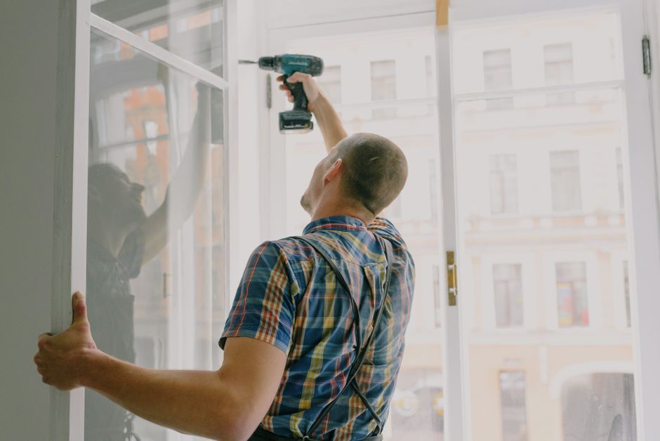 Installer in a plaid shirt drilling into a tall white window frame with an apartment building visible outside