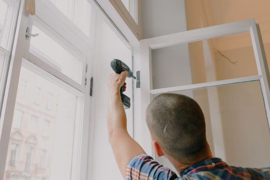 Installer in a plaid shirt using a cordless drill on a white window frame hinge indoors