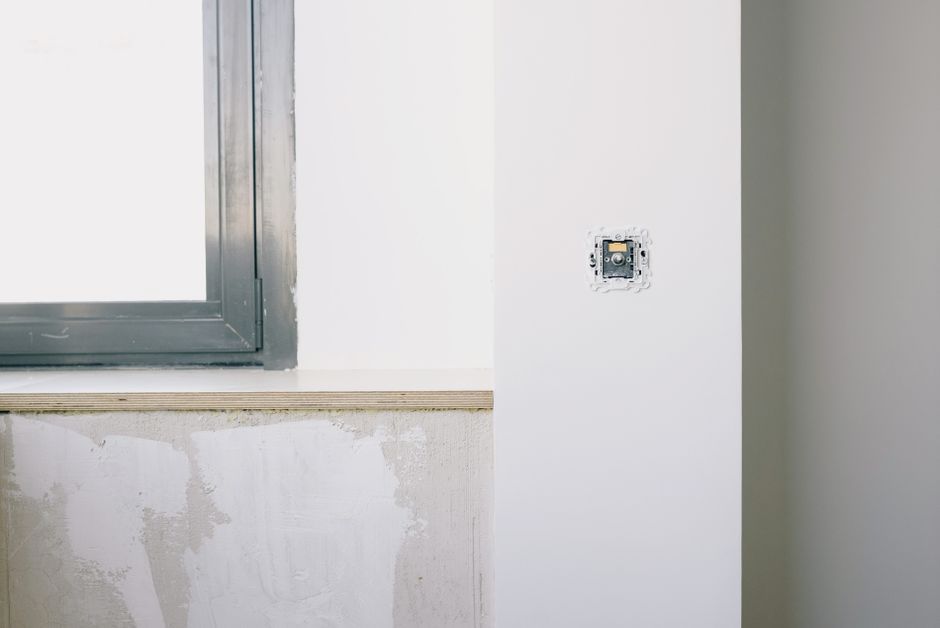 Interior corner with a gray window frame above a plastered sill and an unfinished wall switch socket beside it