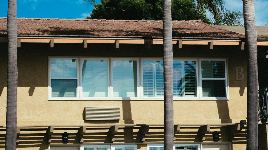 Row of white-framed windows with blinds on a tan stucco building with a tile roof and palm trunks in front