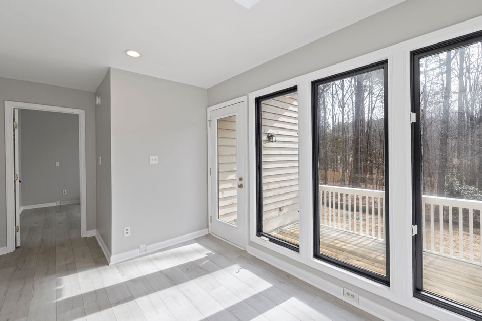 Empty room with light wood floor, a door, and tall black and white framed windows looking onto a wooded deck